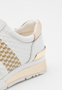 Vit sneaker med strukturerat tyg och metalliska gulddetaljer. Har en tjock sula och mönstrad sida som framhäver varumärkeslogotypen.