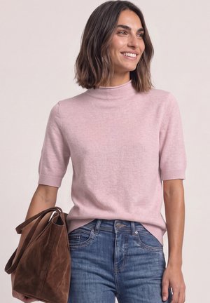 Femme portant un pull léger rose à manches courtes et un jean bleu, souriante, tenant un grand sac à main en daim marron à ses côtés.