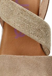Sandal de ante beige con una correa trenzada de acabado texturizado. La plantilla es de cuero marrón, que muestra un logo púrpura tenue.