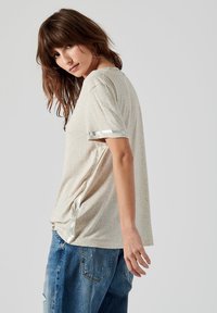 T-shirt beige à manches courtes avec une texture subtile et des accents argentés brillants sur les manches, associé à un jean bleu déchiré.