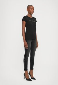 Zwarte aangesloten t-shirt met korte mouwen en gouden scriptlogo, gecombineerd met donkergrijze skinny jeans met zwarte logo-details en zwarte hoge hakken.