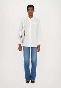 Camicia bianca con colletto a punta e taschino sul petto, abbinata a jeans svasati azzurri chiari e ballerine nere; la modella tiene in mano una piccola borsa color crema.