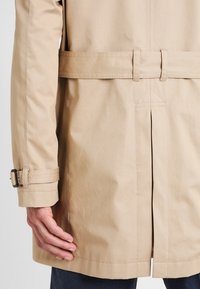 Beżowy płaszcz trench z paskiem w talii, z rozcięciem na plecach i klamrami na rękawach. Gładki, lekki materiał.