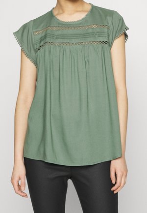 Osoba ubrana w zieloną bluzkę z krótkim rękawem z koronkowym detalem i czarne spodnie, pokazana od szyi do bioder na jednolitym tle.