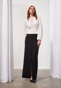 Weiße Bluse mit Spitzenkragen, kombiniert mit einem schwarzen langen Rock mit Schlitz vorne. Glattes Material, taillierter Schnitt und lange Ärmel.
