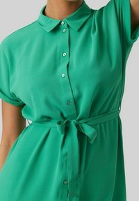 Grön blus med krage, korta ärmar, framknäppning och knut i midjan. Slät tyg med en lätt lyster.