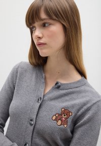 Cárdigan gris hecho de tela suave, con un diseño de botones y un bordado de un oso de peluche en el lado izquierdo del pecho.