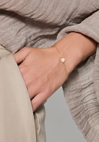 Złota bransoletka z łańcuszka z okrągłym, teksturowanym zawieszką, noszona na dłoni. Bransoletka ma delikatny wzór i subtelny blask w kontraście do beżowego tła.