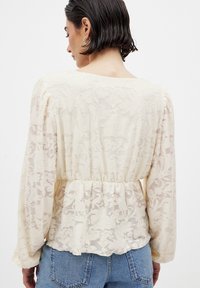 Spitze Peplum-Bluse in hellem Creme mit floralem Muster, langen Ärmeln, elastischem Bund, transparenter Textur und rundem Halsausschnitt.