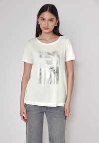 Hvid bomulds t-shirt med en let løs pasform, der har et sølvfarvet grafisk print og tekst i midten. Korte ærmer og rund halsudskæring.