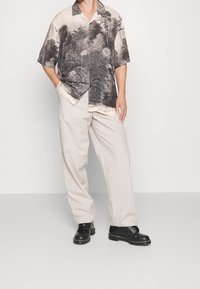 Uomo in pantaloni beige a gamba larga, stivali neri stringati e una camicia a maniche corte con bottoni, decorata con una stampa naturale bianca e nera.