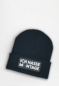 Knit beanie in dark navy with ribbed texture. Features a white rectangular patch with the text "ICH HASSE MONTAGE" and a cat graphic.