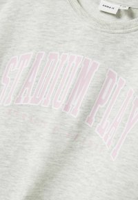 Sudadera gris con cuello acanalado y la inscripción "STADIUM PLAY" impresa en rosa. Textura de tela suave con un diseño casual, ideal para llevar en momentos de relax.