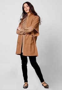 Lichtbruine, gestructureerde cardigan met open voorkant en twee zakken, gelaagd over een tan blouse. Gecombineerd met zwarte slim-fit broeken en platte schoenen.