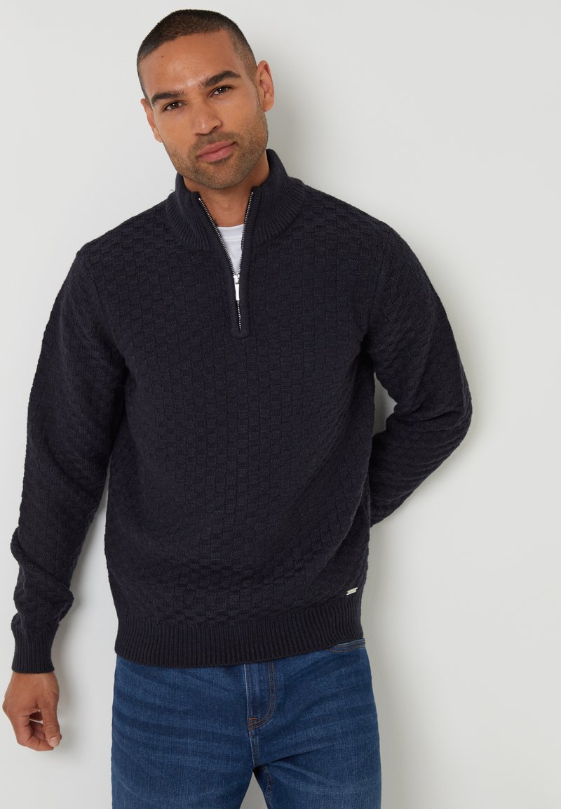 Dunkelblauer Zipperpullover mit strukturiertem Rautenmuster, gerippten Abschlüssen und Saum, getragen über einem weißen Hemd, kombiniert mit blauen Jeans.