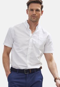 Weißes Kurzarm-Hemd mit Knopfleiste, das über eine Brusttasche, einen Kragen und glattes Baumwollmaterial verfügt, getragen mit einem marineblauen geflochtenen Gürtel.