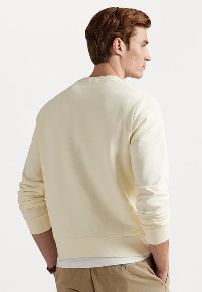 Homem com cabelo castanho, vestindo uma camisa de mangas longas creme e calças bege, de pé com as mãos nos bolsos, de frente para a direita contra um fundo branco.