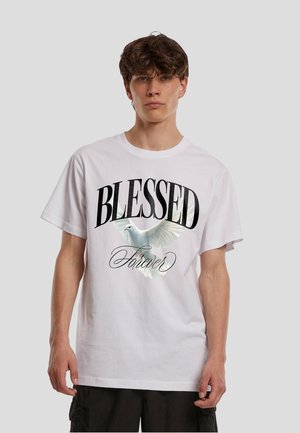Młody mężczyzna z kręconymi włosami, ubrany w białą koszulkę z latającym białym gołębiem i słowami "BLESSED Forever" czarną, skryptową czcionką.