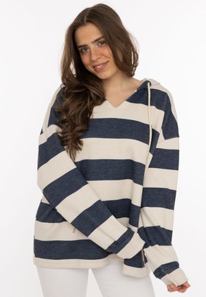 Junge Frau mit langen braunen Haaren, die einen dunkelblauen und beigefarben gestreiften Kapuzenpullover und weiße Hose trägt, lächelt mit verschränkten Händen.