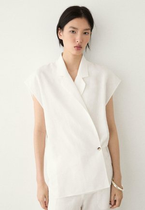 Kvinde med sort hår iført en hvid ærmeløs blazer med en enkelt knap, stående mod en ensartet hvid baggrund og holder et sølvarmbånd.