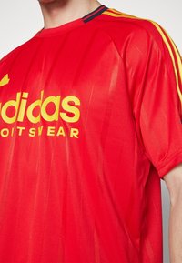 Červené sportovní tričko s žlutým logem "adidas". Obsahuje horizontální pruhy, krátké rukávy a kontrastní modrý límec.