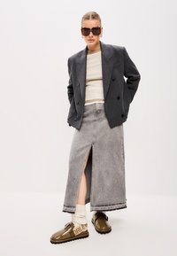 Blazer grigio doppiopetto sopra una maglietta beige, abbinato a una lunga gonna di jeans grigia con spacco frontale e scarpe slip-on marroni con dettagli.