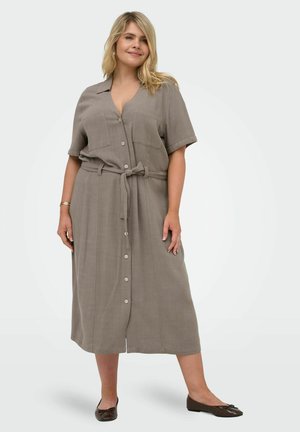 Donna sorridente che indossa un vestito midi con bottoni, maniche corte e cintura e ballerine scure, in piedi davanti a uno sfondo semplice.