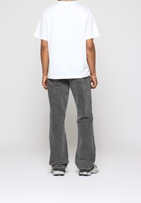 Valkoinen lyhythihainen t-paita ja harmaat leveät housut, joissa on tekstuuripinta. Kengät ja hienovaraiset yksityiskohdat housujen takataskuissa.