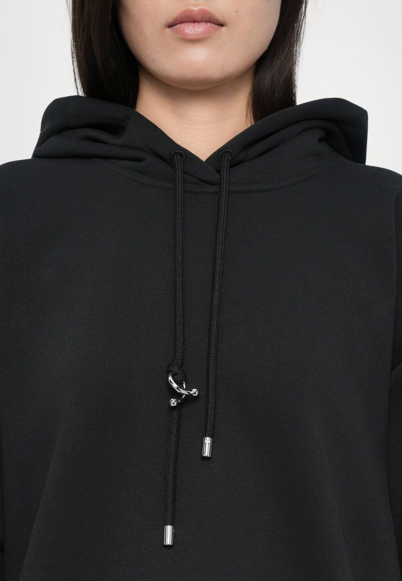 Hoodie noir en tissu mélangé coton, avec un capuchon à cordon de serrage et des embouts argentés. Texture lisse et design minimaliste.