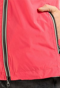 Veste légère rouge avec une texture lisse, dotée d'une poche zippée et d'une fermeture éclair noire contrastante sur le devant.