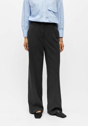 Femme portant un pantalon large gris foncé, une chemise bleu clair à manches longues et des chaussures noires à bout carré, debout devant un fond blanc.