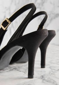 Zwarte suède hoge hakken sandaal met een taps toelopende hak, voorzien van slanke bandjes en een gouden gespdetail. Textureafwerking en een gladde afwerking.