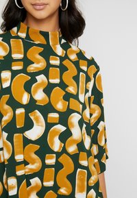 Zelená blůza s abstraktními malířskými vzory v oranžové a bílé barvě, vysokým výstřihem a 3/4 rukávy, vyrobená z měkké texturované látky.