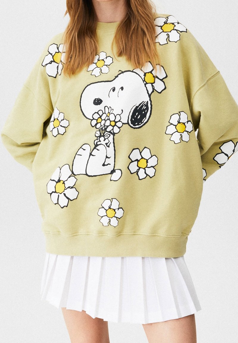 Femme portant un sweat-shirt vert clair avec Snoopy tenant des fleurs et des marguerites blanches partout, associé à une jupe plissée blanche.