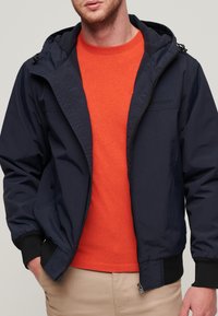 Marineblå hættejakke med frontlynlås og elastiske manchetter. Bæres over en orange t-shirt med rund hals. Glat stof, minimal branding.