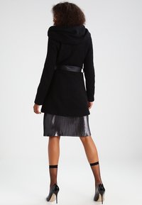 Cappotto lungo nero con cappuccio, cintura in vita, abbinato a una gonna plissettata argento e collant in rete nera, indossato con tacchi alti neri.