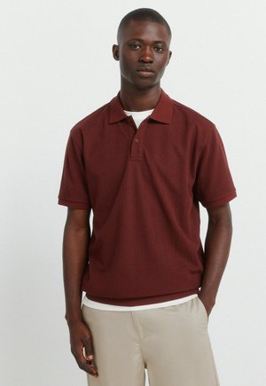 Junger Mann steht mit den Händen in den Taschen, trägt ein kastanienbraunes kurzärmeliges Poloshirt über einem weißen Unterhemd und beigen Hosen vor einem schlichten Hintergrund.