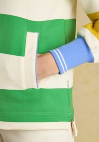 Hand in grün-weiß gestreifter Tasche mit blauem Manschettenbund und gelbem Ärmel auf cremefarbenem Hintergrund sichtbar.