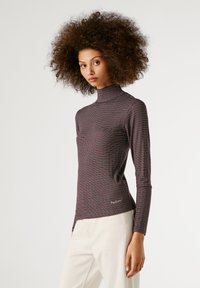 Noor nooriga volüümsete lokkis juustega, kandes triibulist kõrge kaelusega pluusi ja valgeid pükse, seistes ühetoonilise heleda tausta ees.