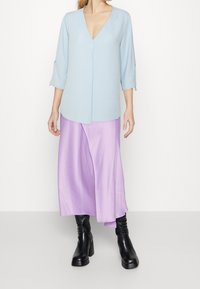 Blusa di un blu chiaro, con texture, scollatura a V e maniche a tre quarti, abbinata a una gonna fluida in satin lavanda e stivali neri fino al ginocchio.