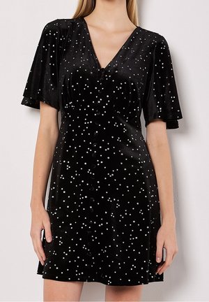 Zwart fluwelen korte jurk met zwierige mouwen, V-hals, knopen aan de voorkant en een verspreid wit sterrenpatroon, gedragen door een staande persoon.