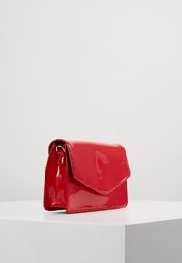 Bolso de mano de charol rojo con cierre de solapa, forma rectangular y una textura suave y brillante. Presenta un asa lateral.