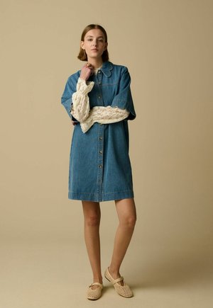 Femme portant une robe en denim longueur genoux avec col festonné et manches en dentelle, debout les bras croisés et chaussures tissées beiges sur fond beige.