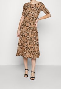 Kvinna klädd i en brun och beige blommig paisleymidiklänning med ärmar som går till armbågen och svarta sandaler med remmar runt anklarna, stående med en hand på höften.