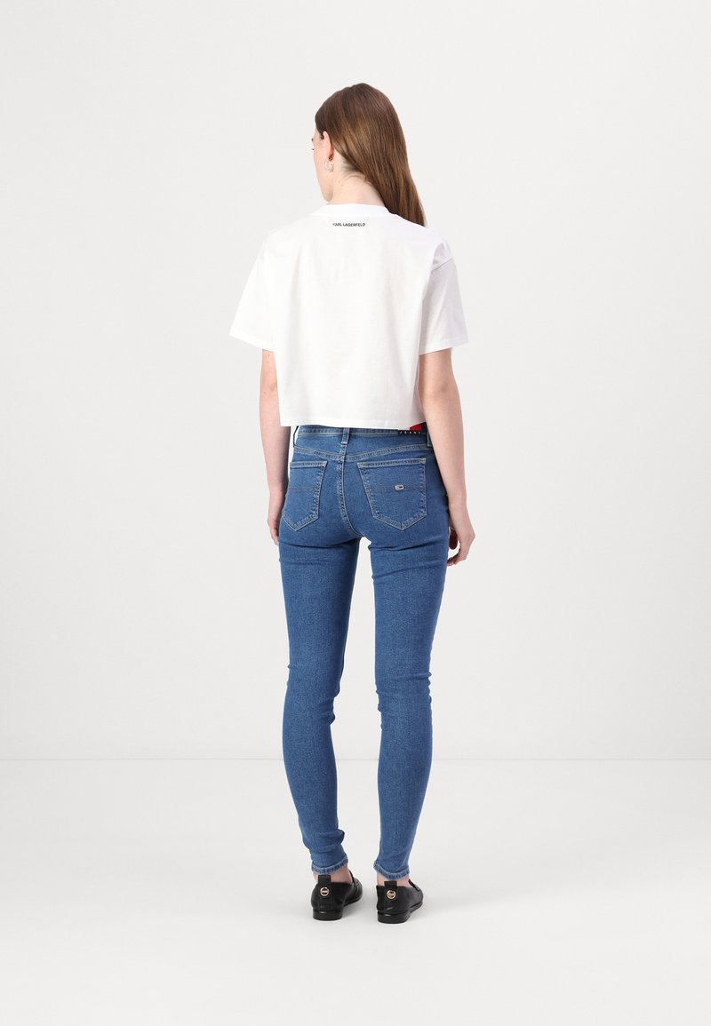 Camiseta blanca corta con mangas cortas, con un logo en la parte posterior. Jeans ajustados azules con cinco bolsillos y un ligero estiramiento. Zapatos negros.