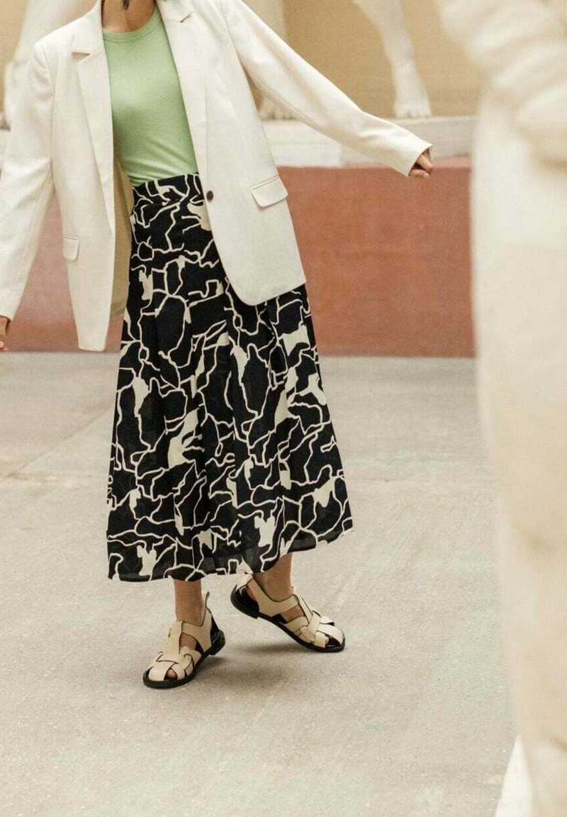 Personne portant un blazer clair, un top vert, une jupe à motifs noir et blanc et des sandales beige, debout sur une surface en béton.