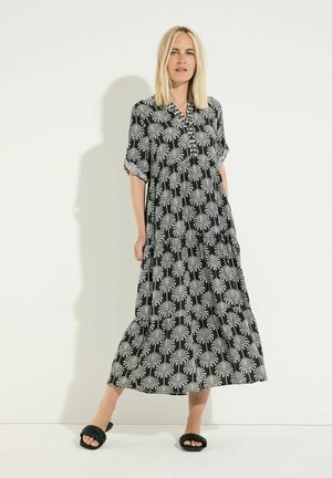 Blondine draagt een lange zwart-wit palmboomjurk met opgerolde mouwen en zwarte geweven instapsandalen.