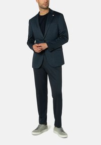 Op maat gemaakte donkerblauwe colbert met bijpassende broek, vervaardigd uit een gestructureerde stof. Gecombineerd met lichtgrijze sneakers voor een moderne uitstraling.