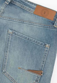 Jeans de denim en azul claro con un parche de cuero y dos bolsillos traseros; costuras sutiles y un pequeño detalle de bolsillo con cremallera.