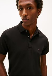 Mladý muž s krátkými kudrnatými vlasy, oblečený v černém polotriku se třemi knoflíky a malým červeno-bílo-modrým logem na hrudi.